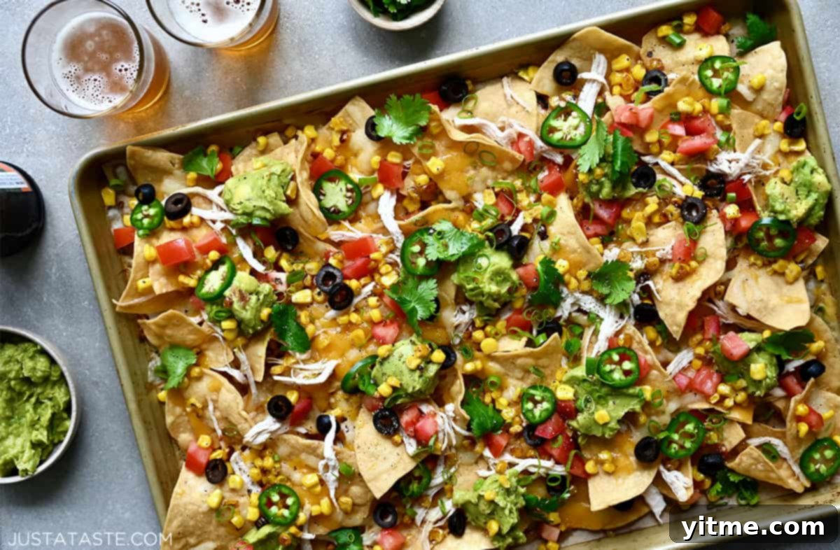 A top-down view of sheet pan nachos loaded with sliced jalapeños, black olives, corn kernels, diced tomatoes, shredded cheese and shredded chicken.