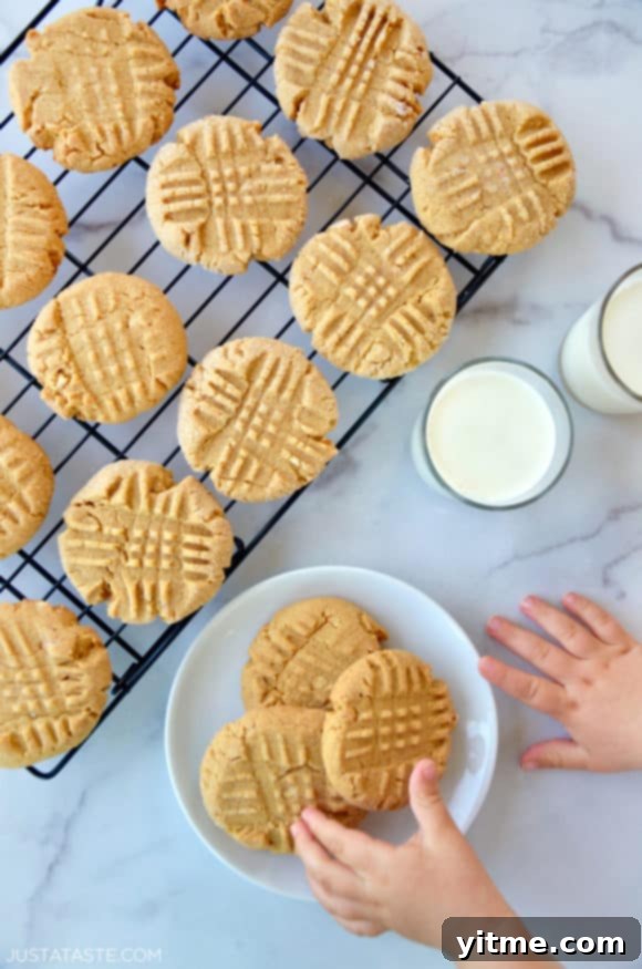 Child's hand reaching for Soft and Chewy Peanut Butter Cookies on white plate next to glasses of milk and wire cooling rack with cookies