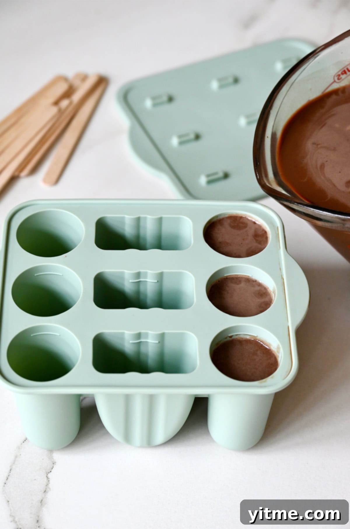 Pouring homemade fudgesicle mixture from a liquid measuring cup into flexible silicone popsicle molds.