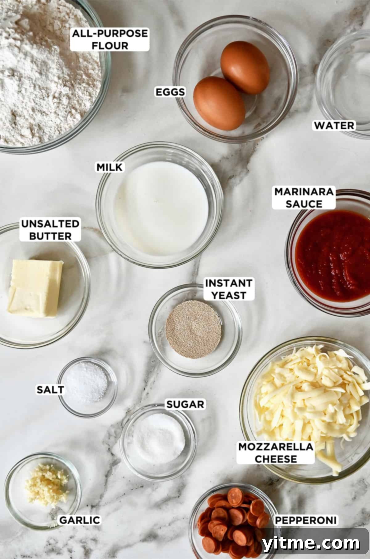 Various sizes of clear bowls containing flour, eggs, milk, marinara sauce, instant yeast, shredded mozzarella cheese, salt, sugar, butter and mini pepperoni.