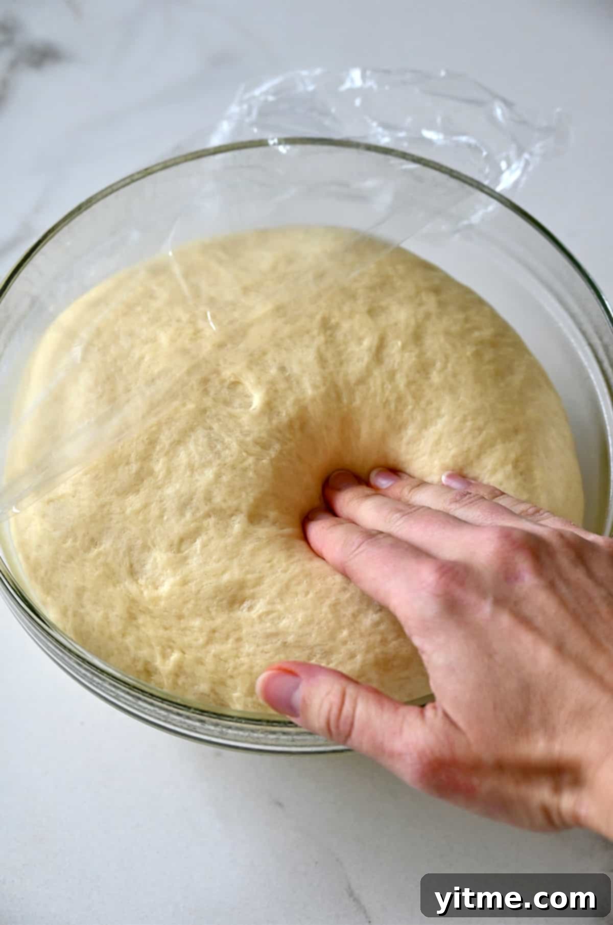 A hand presses into proofed dough in a glass bowl.