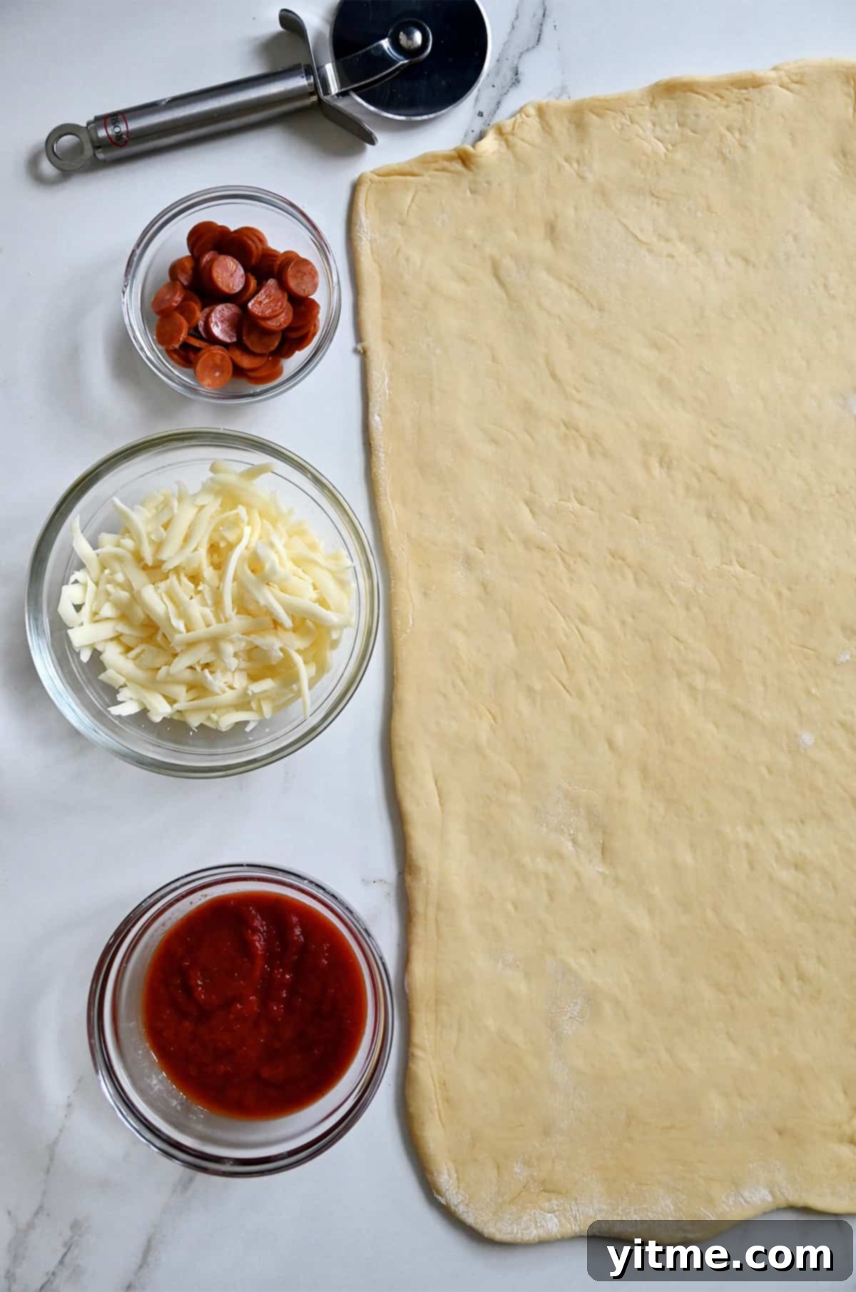 Dough rolled out into a rectangle next to small bowls containing marinara sauce, shredded cheese and mini pepperonis.