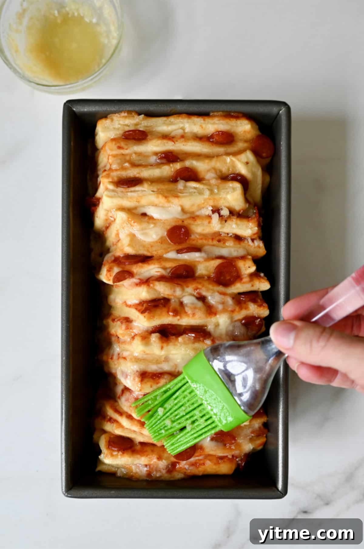 A hand holding a pastry brush applies garlic butter atop unbaked pizza bread in a loaf pan.
