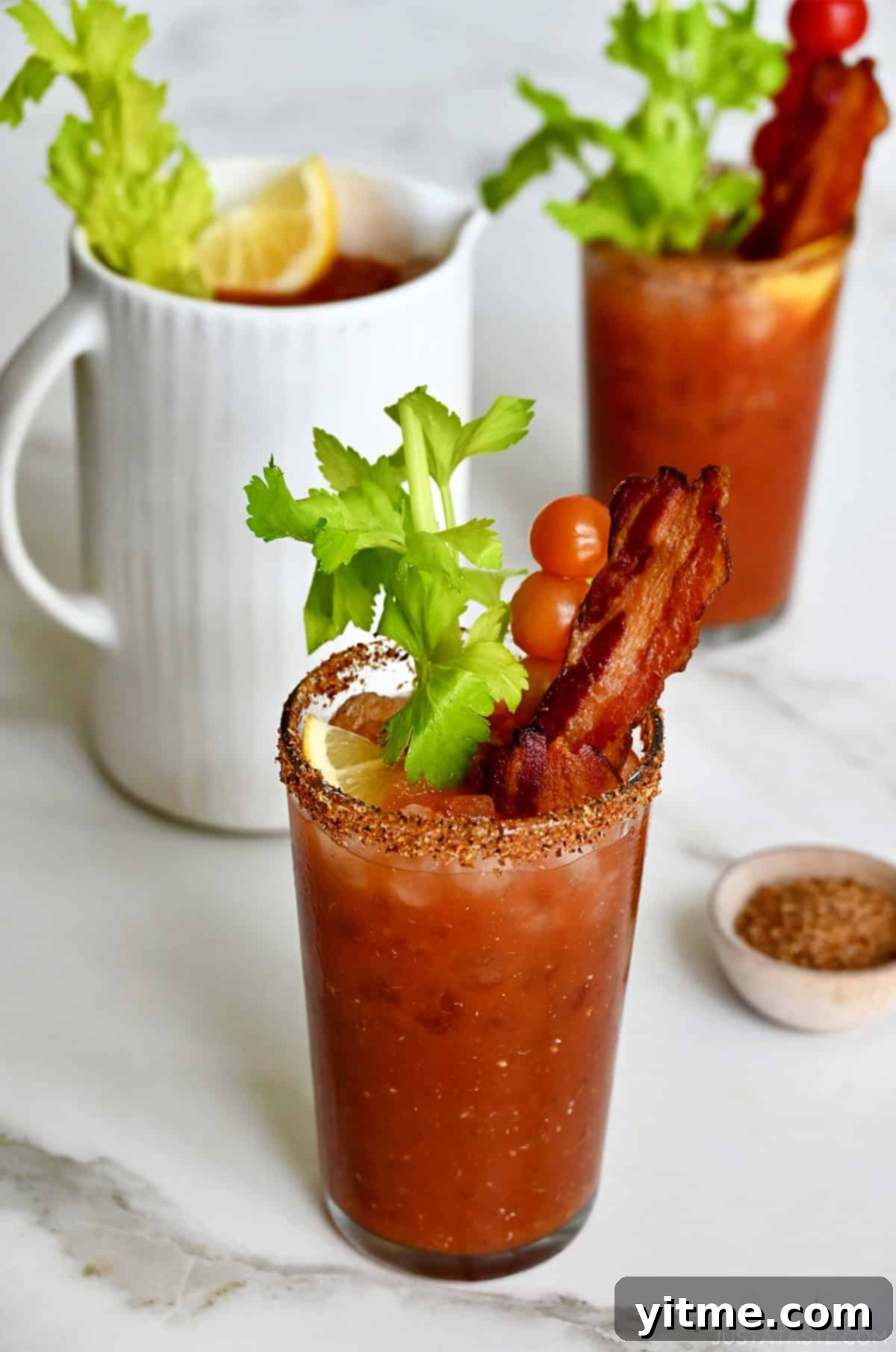 A pitcher of Bloody Marys next to two glasses rimmed with Tajin, filled with Bloody Mary mix and garnished with a celery stalk, a skewer of cherry tomatoes and crispy bacon.