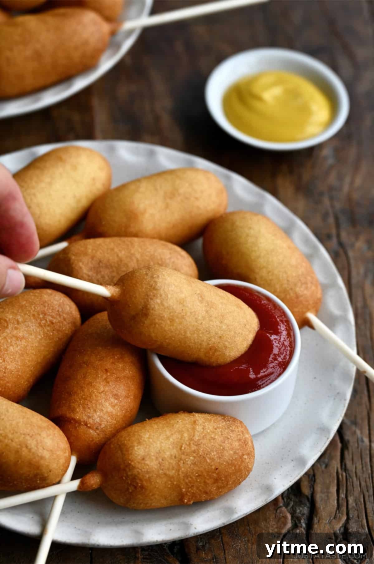 A hand dips a homemade mini corn dog on a stick into a small bowl of ketchup.