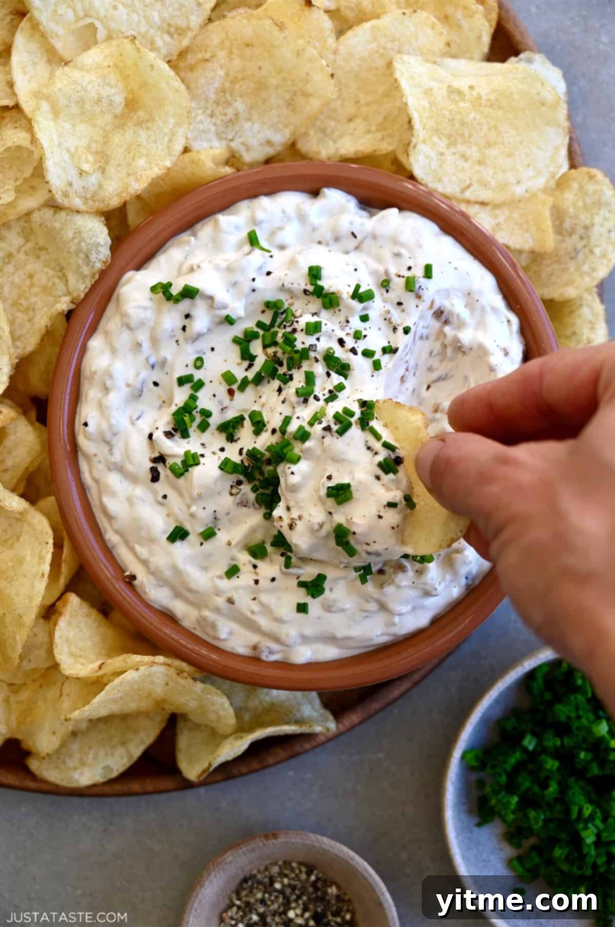 A hand holding a potato chip dips it into a bowl containing sour cream and onion dip that's on a plate surrounded by potato chips.