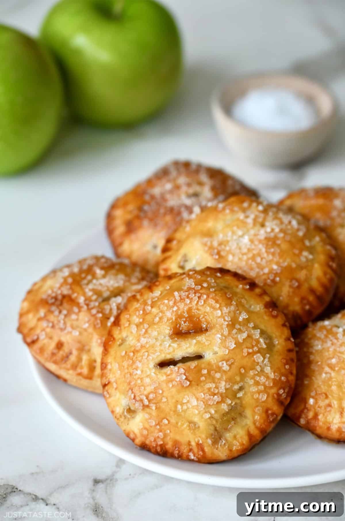 Golden-brown salted caramel apple hand pies with a sparkling sugar topping, arranged on a white plate with fresh Granny Smith apples in the background, hinting at the crisp, tart apples within.