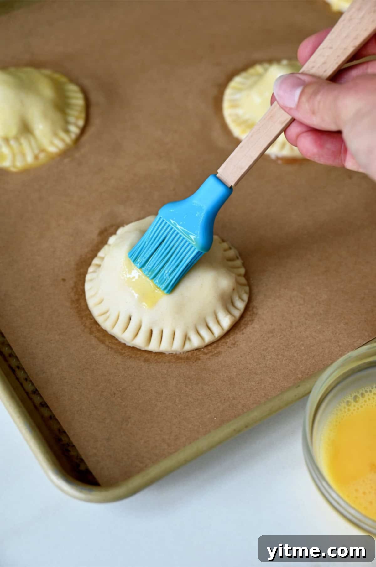 A hand using a pastry brush to apply an egg wash over unbaked hand pies resting on a parchment paper-lined baking sheet, preparing them for a golden crust.