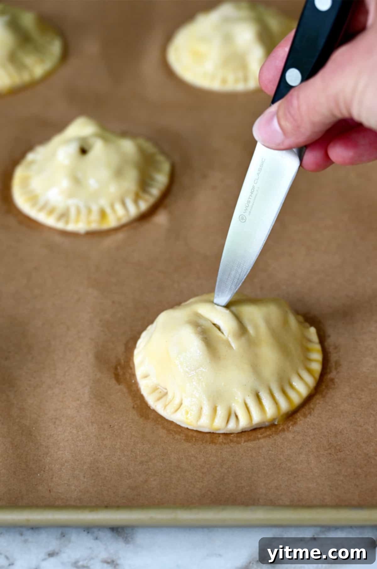 A hand holding a sharp knife carefully cuts slits into the tops of unbaked apple hand pies on a parchment-lined baking sheet, allowing steam to escape during baking.