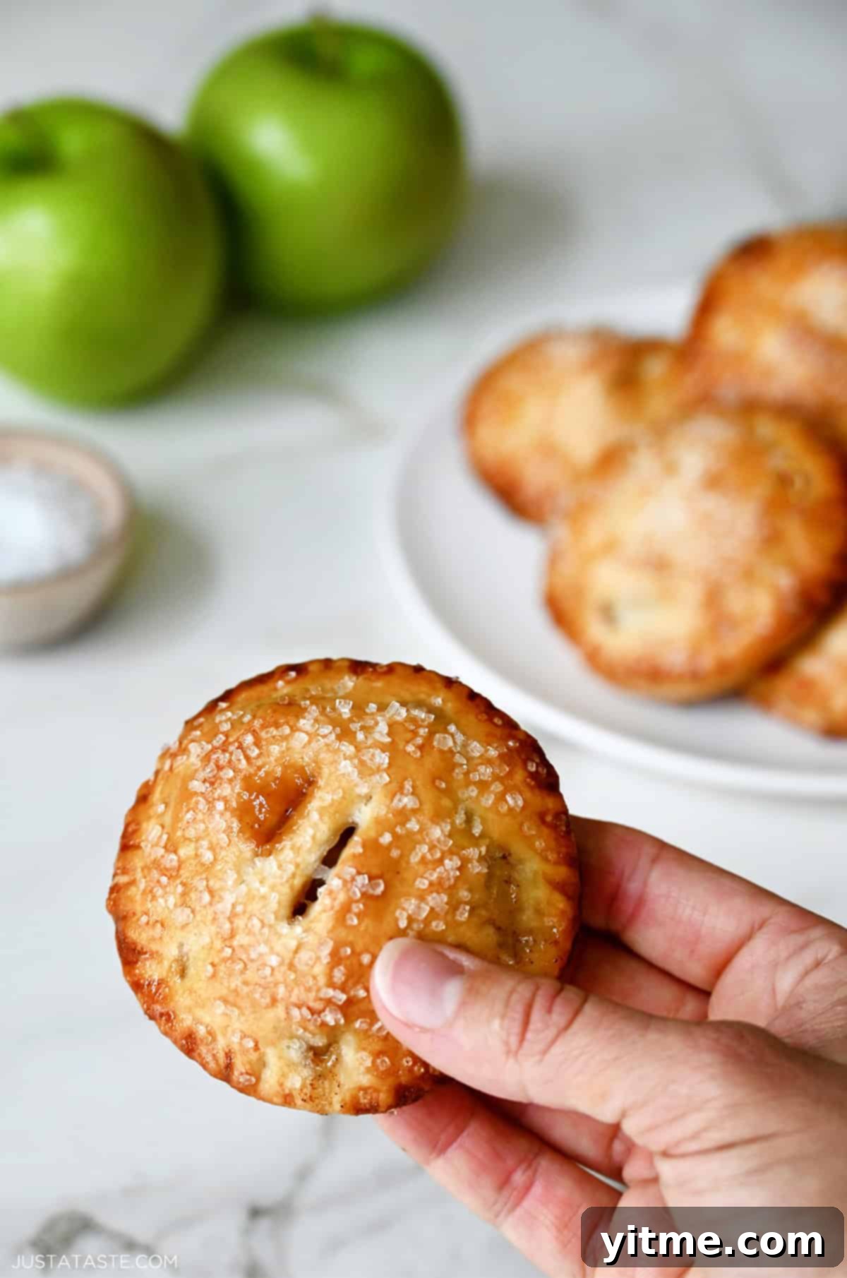 A hand delicately holds a single, perfectly golden-brown mini apple hand pie, showcasing its individual portion size and inviting appearance.