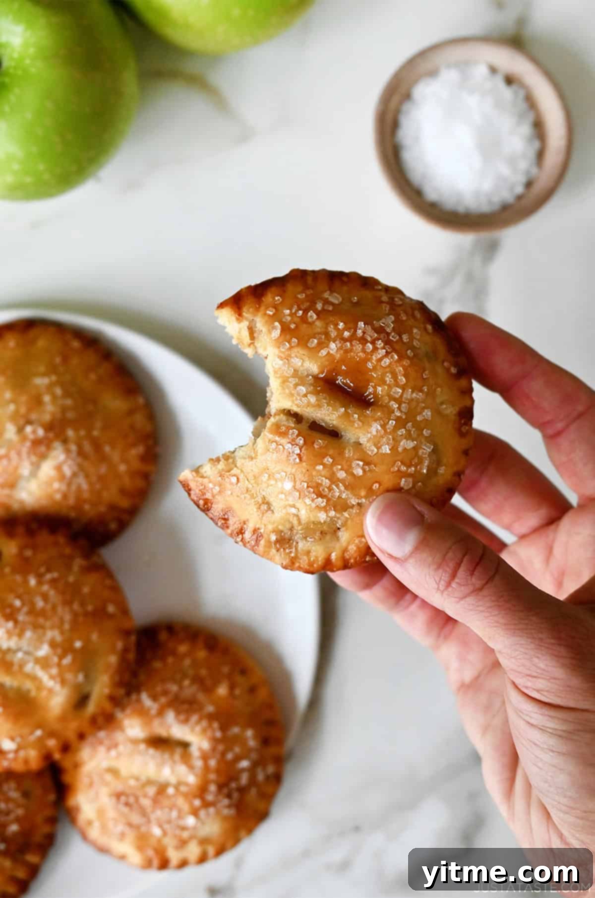 A hand holds a freshly baked salted caramel apple hand pie with a bite taken out, revealing the soft apple and gooey caramel filling inside.