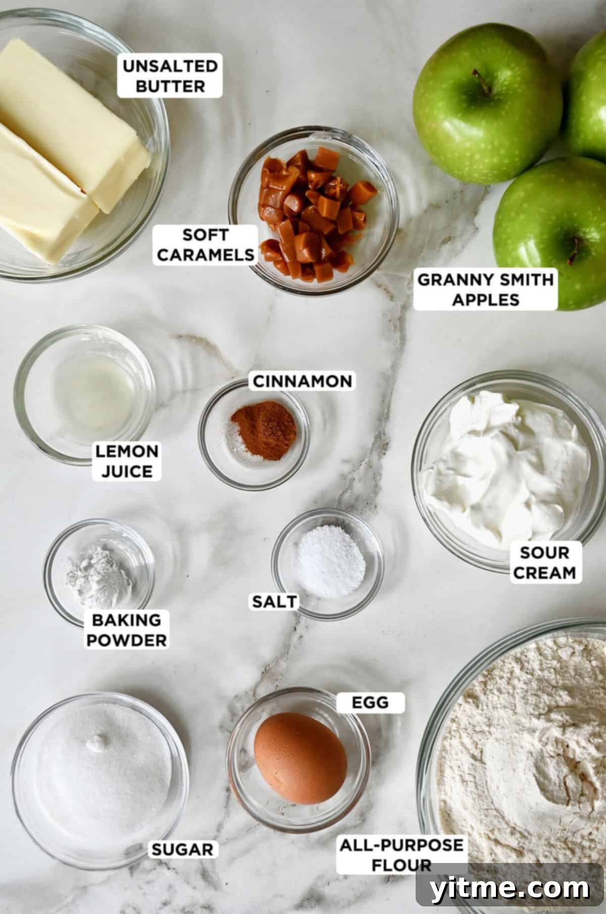 Various ingredients for salted caramel apple hand pies laid out in glass bowls: two green Granny Smith apples, sour cream, flour, an egg, granulated sugar, baking powder, fresh lemon juice, ground cinnamon, cold butter cubes, and soft caramel candies.