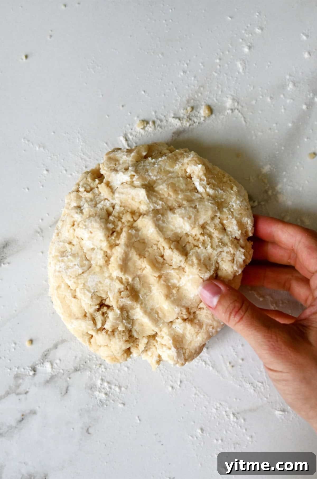 A hand gathering shaggy, crumbly pie dough on a flour-dusted marble surface, illustrating the initial stage of bringing the dough together.