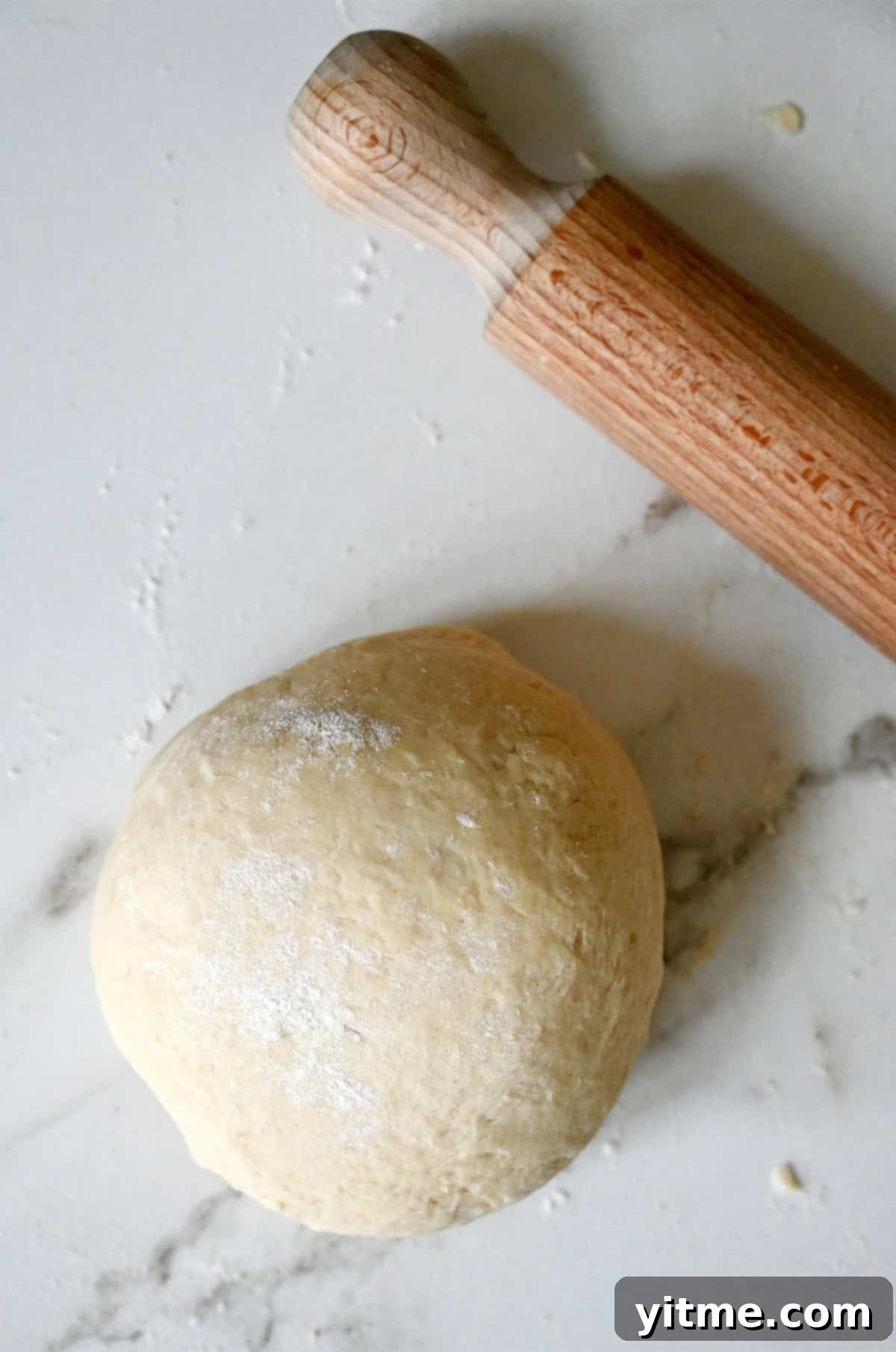 Smooth, cohesive pie dough lightly dusted with flour resting on a polished marble surface, ready for rolling, with a wooden rolling pin nearby.