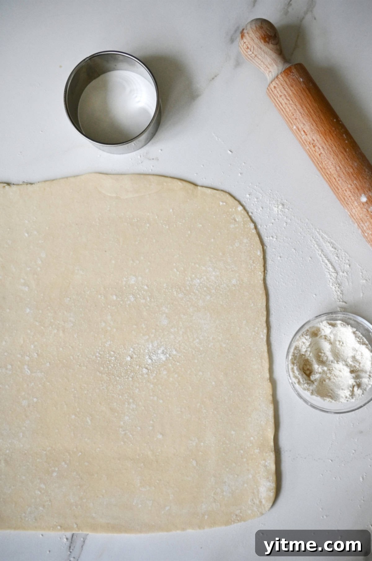 Neatly rolled out pie dough on a flour-dusted surface, accompanied by a wooden rolling pin, a small bowl of flour for dusting, and a round cookie cutter, all set for pie assembly.