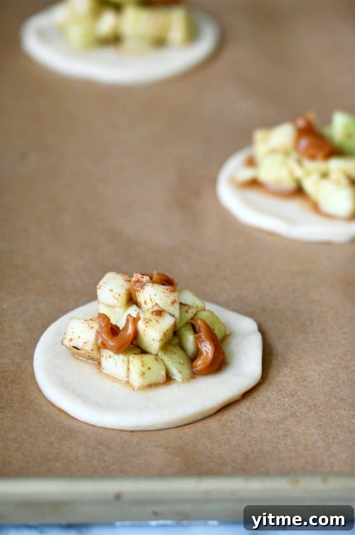 A baking sheet lined with parchment paper, showcasing circular pie dough bases topped with generous spoonfuls of caramel apple pie filling, ready for the top crust.