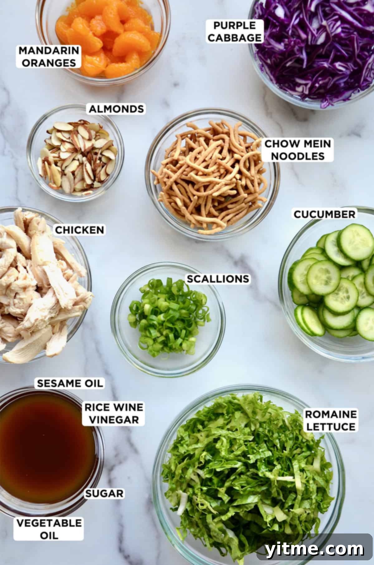 Chinese chicken salad ingredients in various sizes of glass bowls, including mandarin oranges, shredded purple cabbage, crispy chow mein noodles, sliced cucumbers, chopped scallions, shredded romaine lettuce, sesame dressing, cooked chicken breast slices and sliced almonds.
