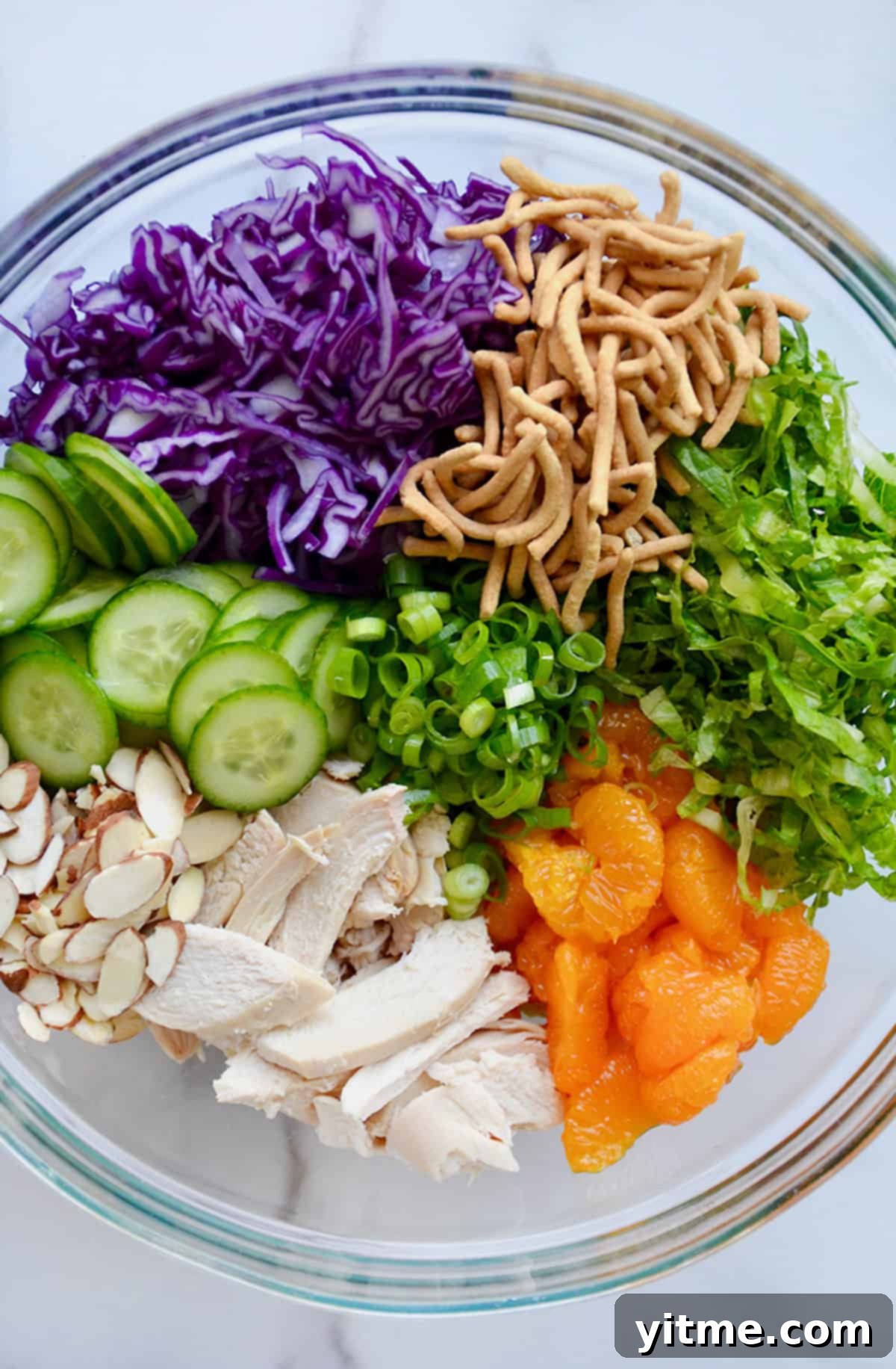 Shredded chicken breast, sliced almonds, sliced cucumbers, shredded purple cabbage, chopped scallions, crispy chow main noodles, shredded romaine lettuce and mandarin oranges in a large glass bowl.