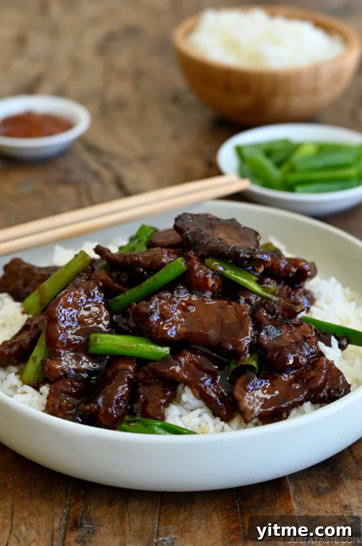 Mongolian beef with sliced scallions atop steamed white rice in a white bowl with chopsticks.