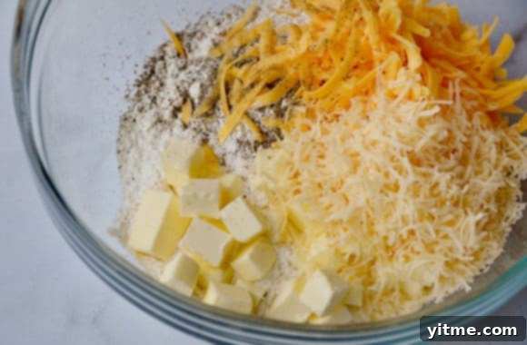 Ingredients for cheddar biscuits laid out in a glass bowl: flour, cold butter cubes, and a generous heap of shredded cheddar cheese, emphasizing fresh preparation.
