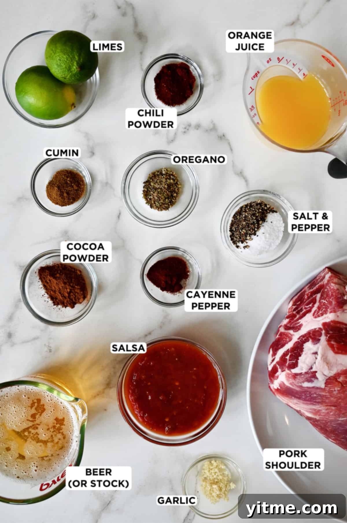 Small bowls of limes, chili powder, oregano, cumin, salt and pepper, cocoa powder, cayenne pepper, salsa and minced garlic on a marble countertop. Measuring cups containing beer and orange juice and plate with a pork shoulder on it are also on the countertop.