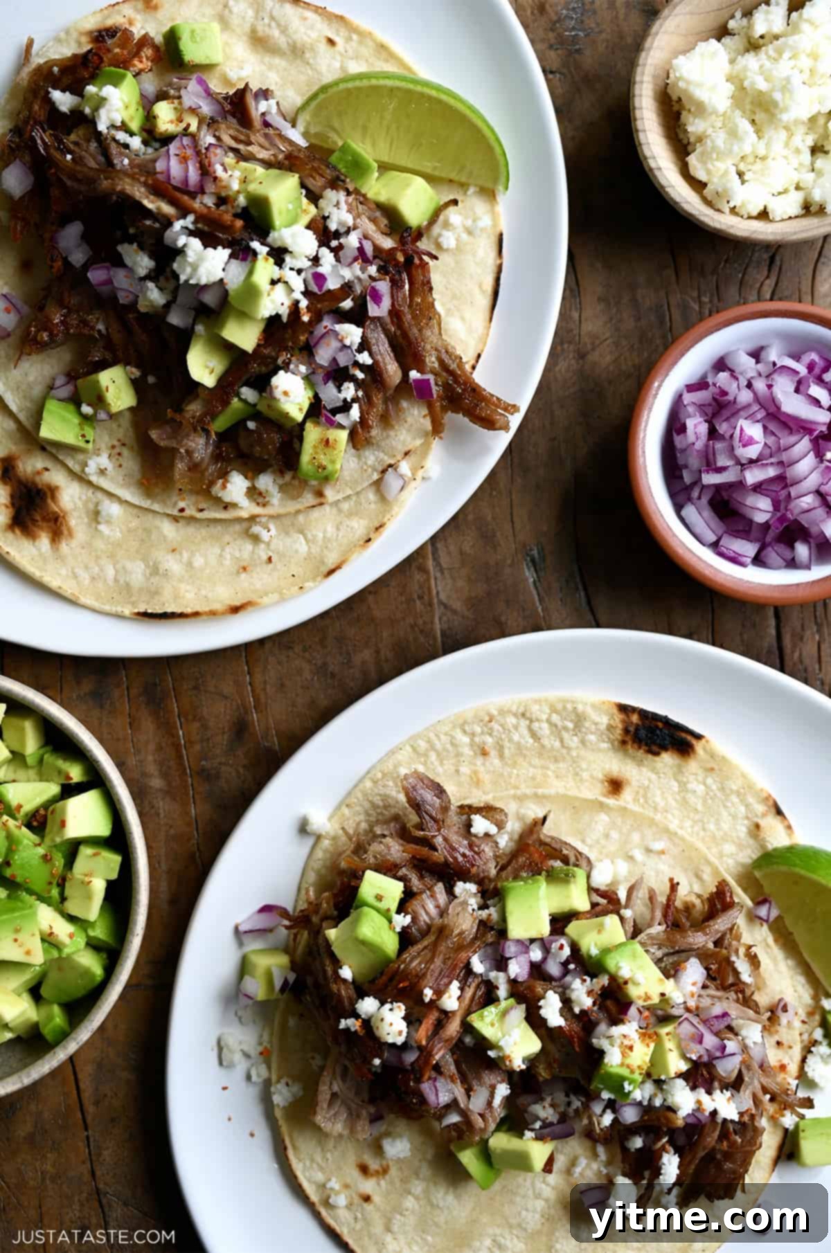 Two pulled pork tacos on separate plates. The tacos are topped with cubed avocado, crumbled cotija cheese and diced red onion. Small bowls with more toppings are beside the tacos.