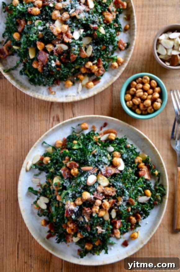 Two white plates piled high with vibrant kale salad, generously topped with warm bacon vinaigrette, crispy chickpeas, and toasted nuts, with a small bowl of extra crispy chickpeas on the side