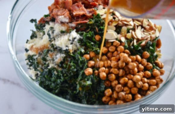 Close-up view of warm bacon dressing being poured from a small saucepan onto a glass bowl filled with colorful kale salad ingredients, demonstrating the crucial dressing step