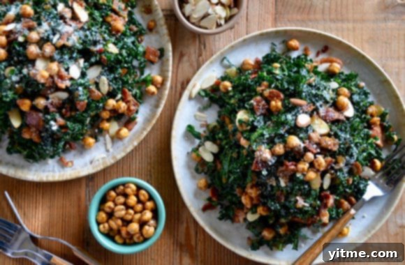 Two pristine white plates elegantly arranged with servings of fresh kale salad, featuring a generous drizzle of warm bacon vinaigrette and accompanied by small, inviting bowls of extra crispy chickpeas and toasted almonds