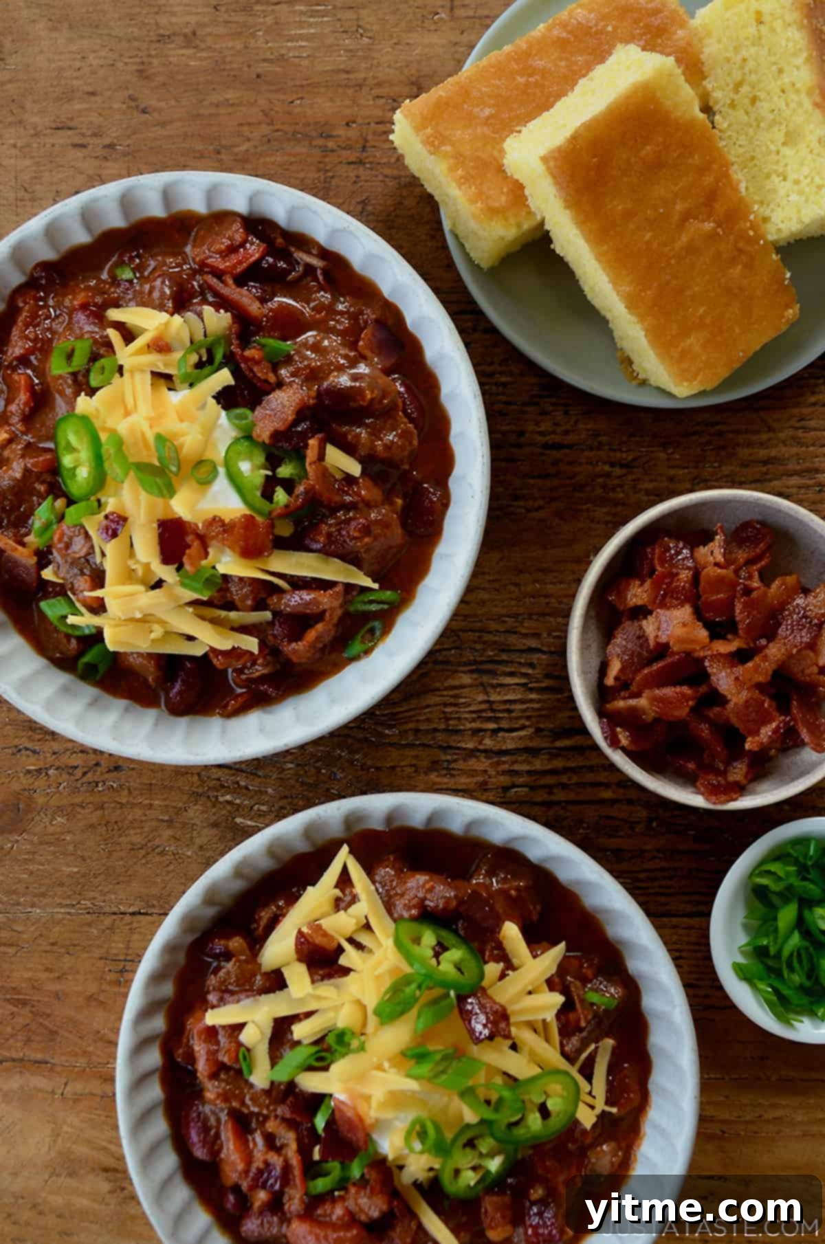 Bowls of Chipotle Chili con Carne topped with sour cream, bacon, cheese, jalapenos, and scallions with cornbread on the side