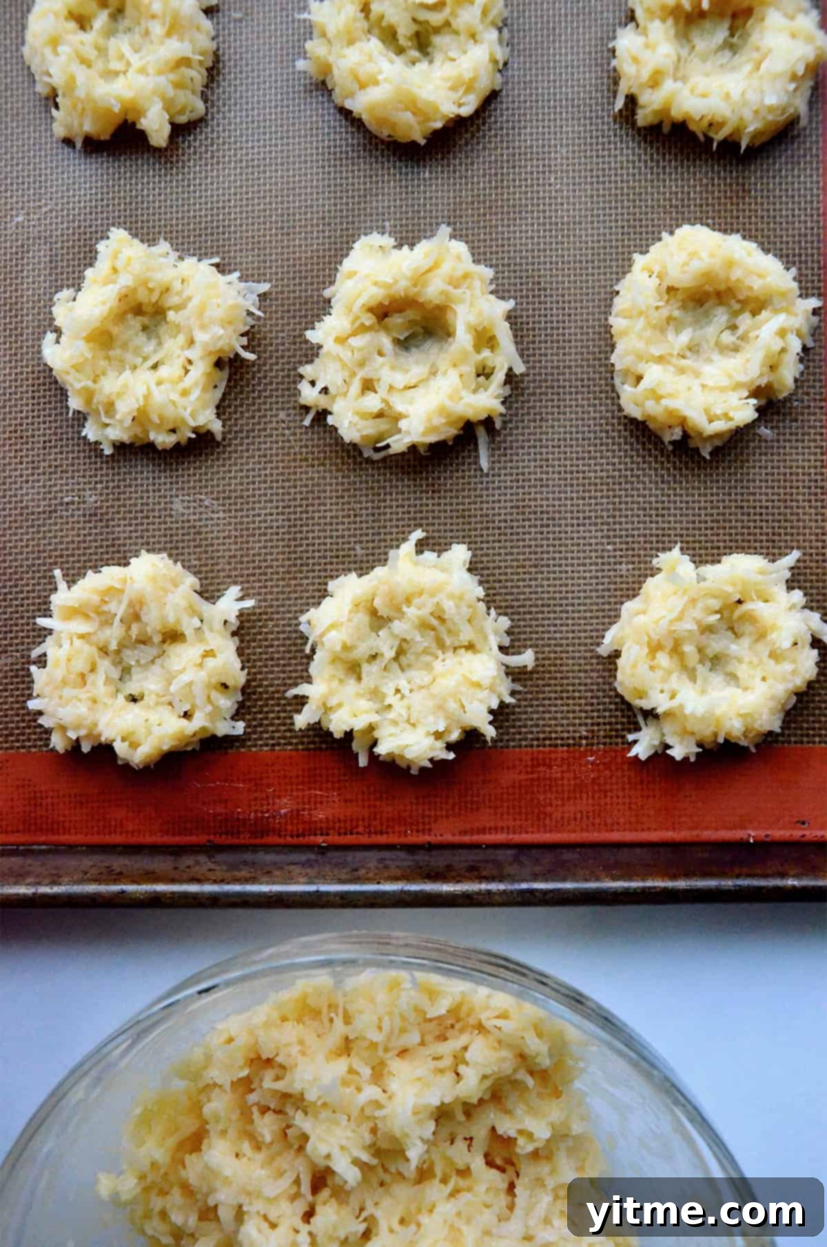 Coconut macaroon nests are lined up on a silpat baking sheet, ready to be baked. A bowl of macaroon batter in a glass bowl is nearby.