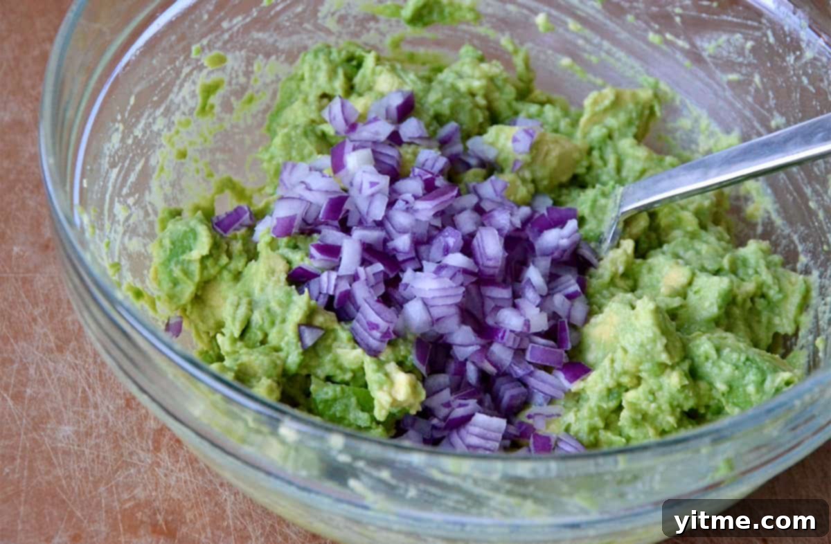 Diced red onion on top of mashed avocado in a glass mixing bowl.