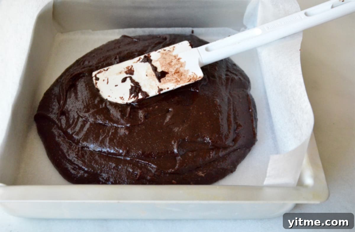 Brownie batter in a square baking dish lined with parchment paper. A rubber spatula rests against the side of the dish.