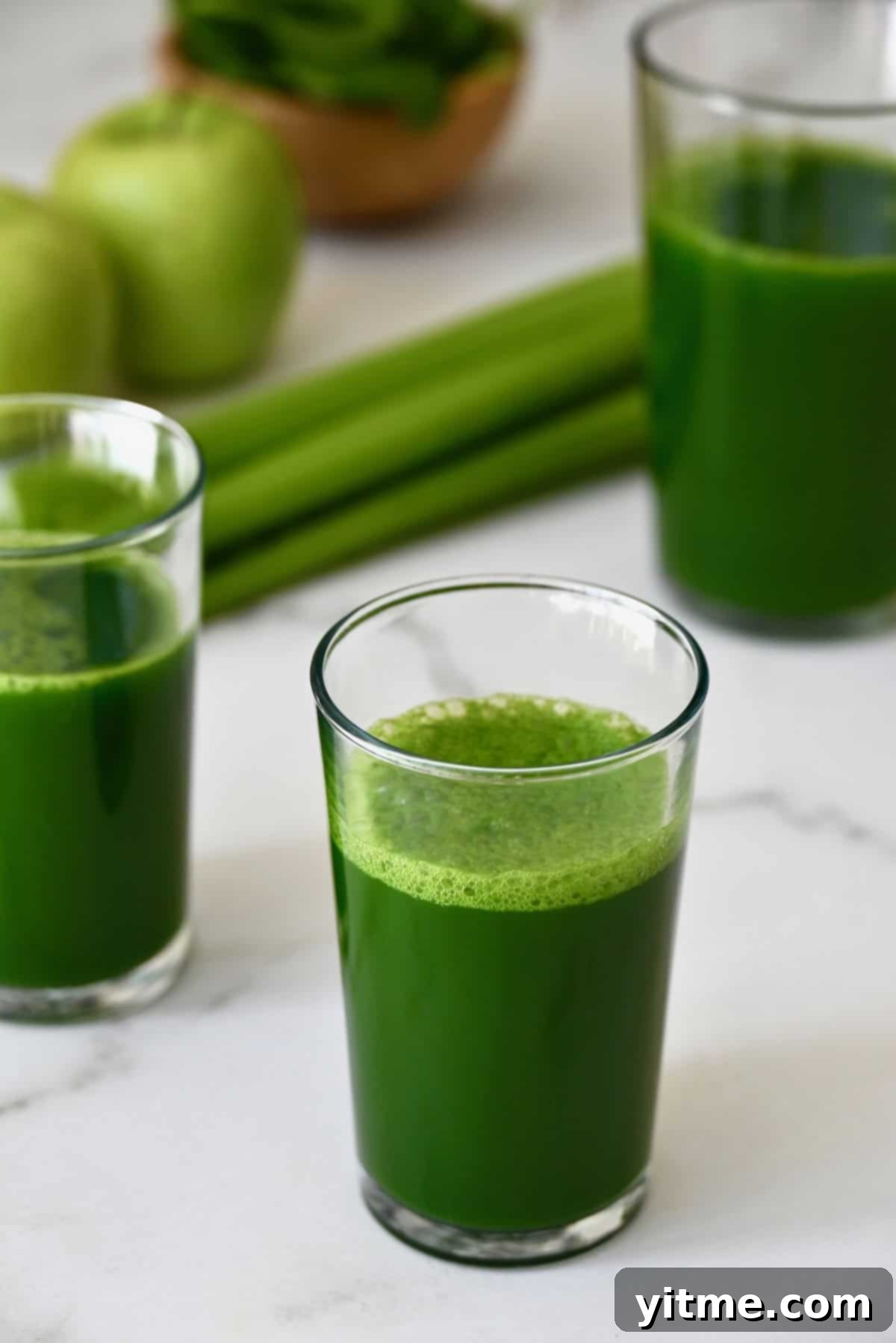 Glasses of green juice sit on a white marble countertop with celery and apples in the background.