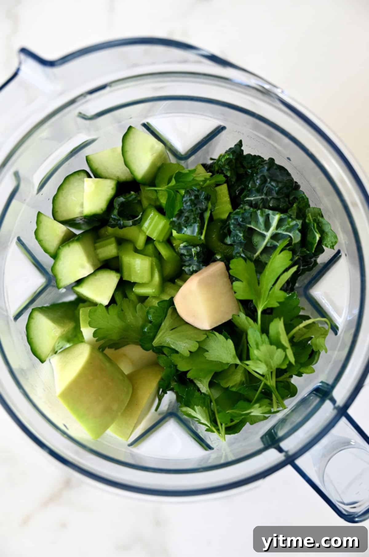 A blender jar is filled with ingredients for green juice, including cucumber, celery, ginger, apple and kale.