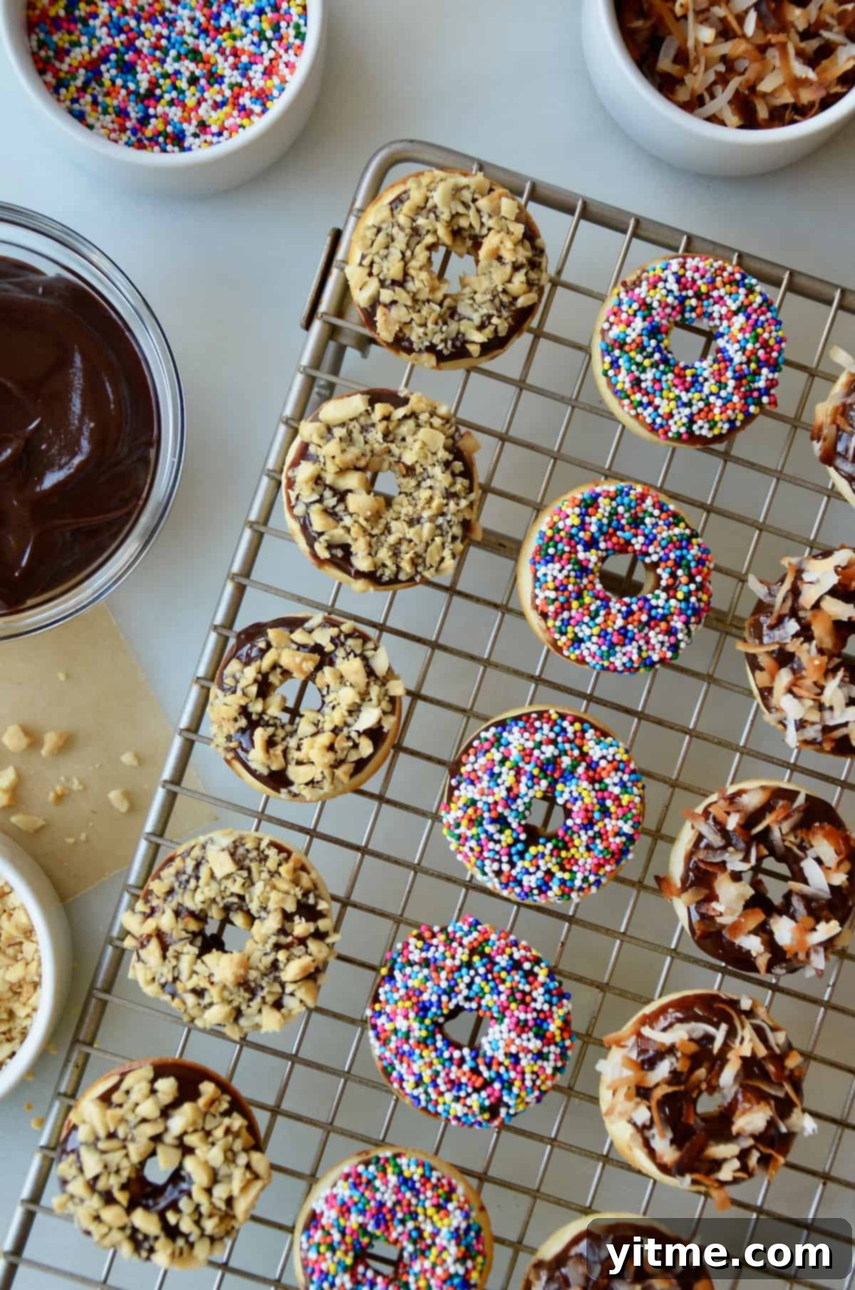A cooling rack with baked and glazed doughnuts topped with chopped nuts, sprinkles, and toasted coconut. Bowls with more toppings and Nutella glaze are beside the cooling rack.
