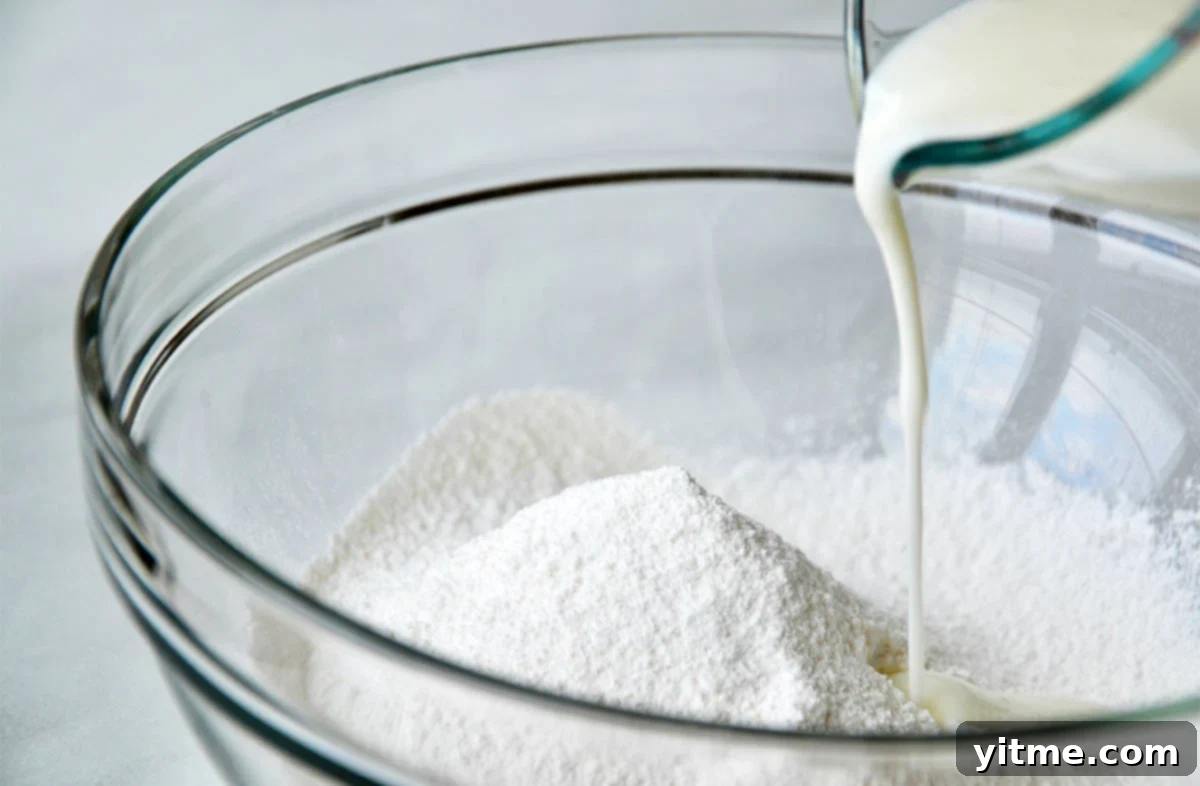 Measuring cup pouring liquid ingredients into a flour mixture in a large glass mixing bowl.