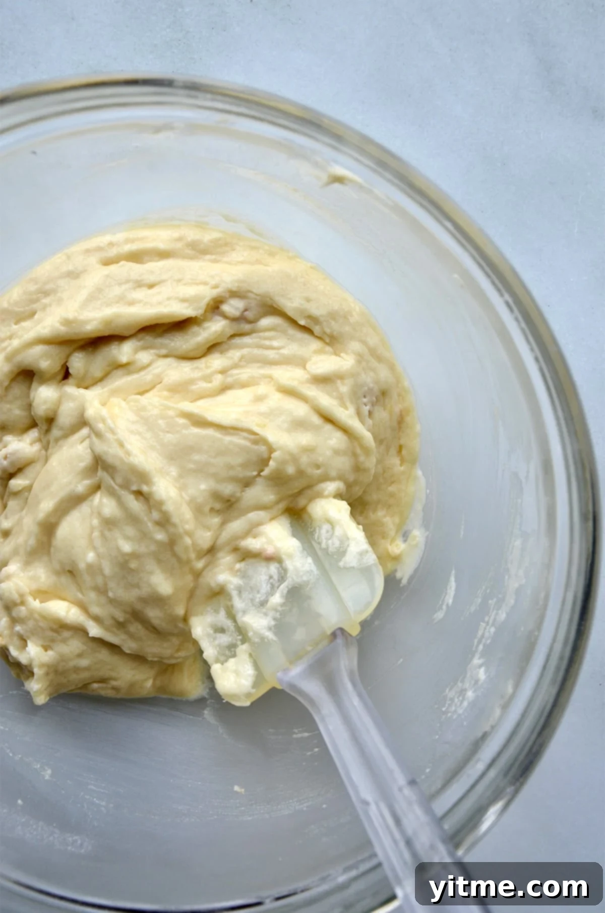 A large glass mixing bowl with doughnut batter in it. A spatula rests in the batter and against the side of the bowl.