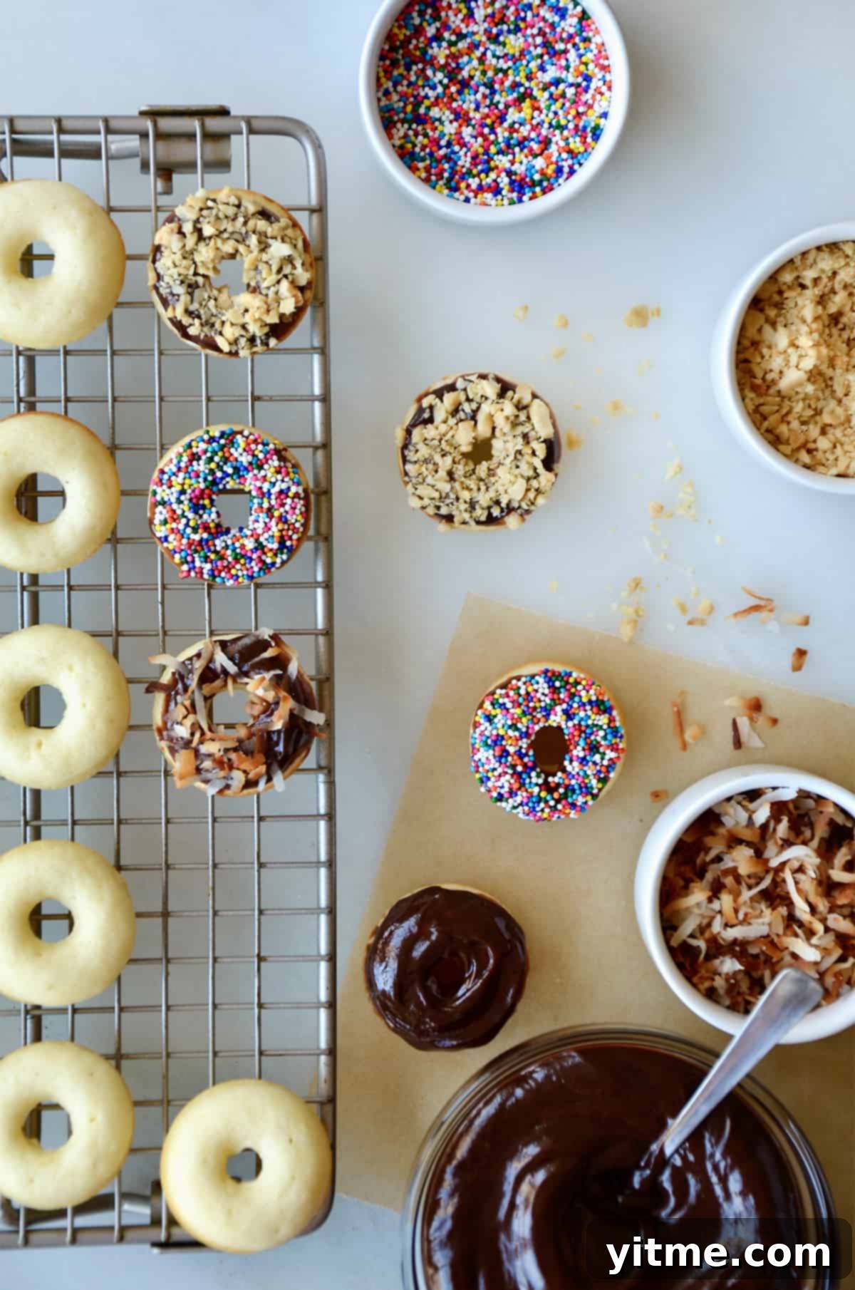 Baked doughnuts on a wire cooling rack. Topped doughnuts and bowls of Nutella glaze, sprinkles, chopped nuts and toasted coconut are beside the cooling rack.