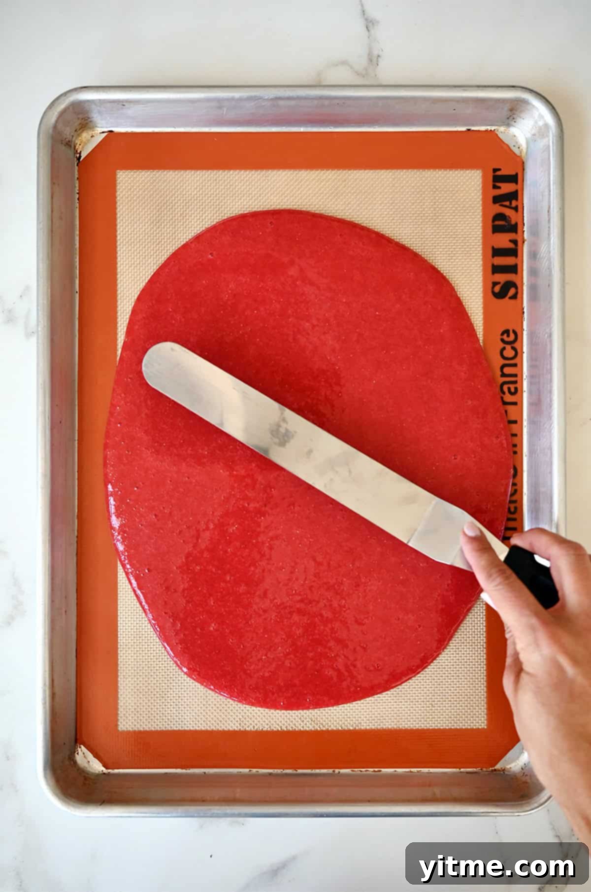 A hand holding an offset spatula spreads strawberry purée atop a baking sheet lined with a silicone baking mat.