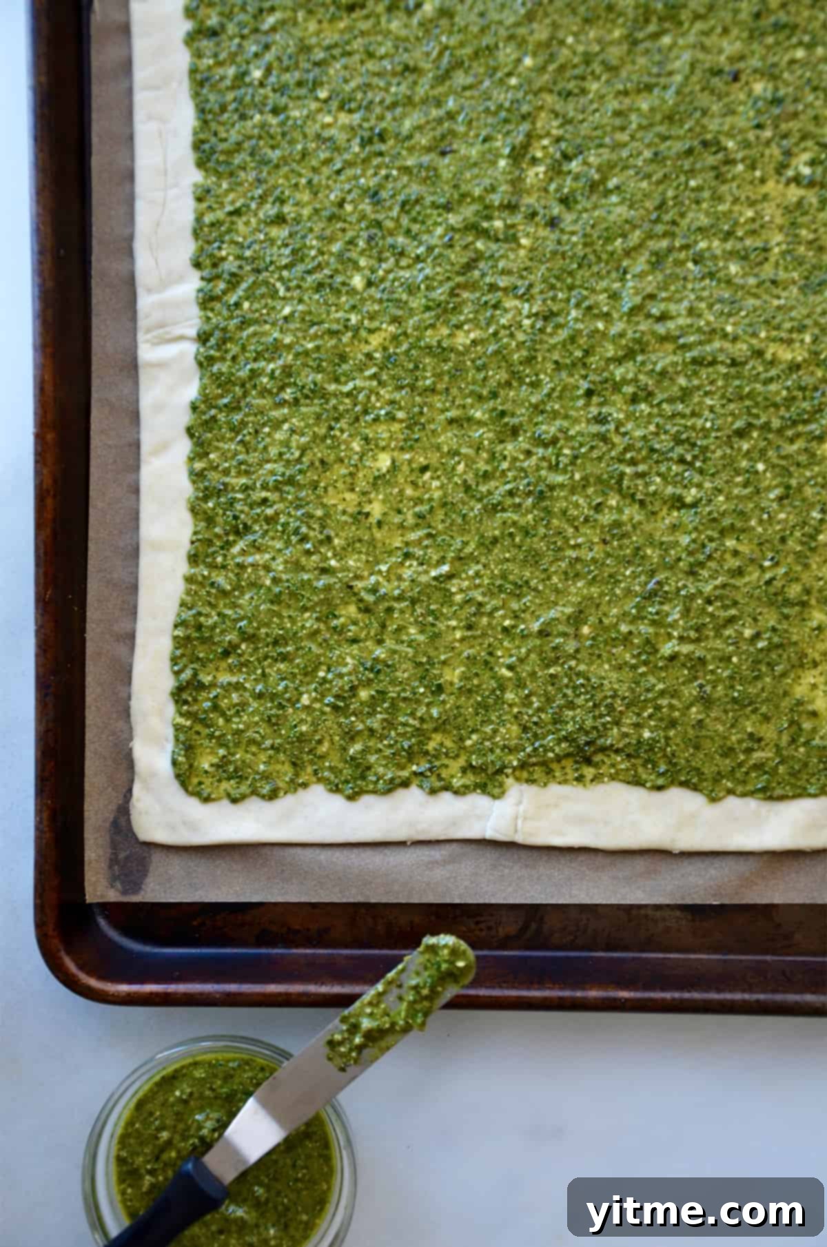 A rectangle of puff pastry topped with pesto and placed on a rimmed baking sheet lined with parchment paper.