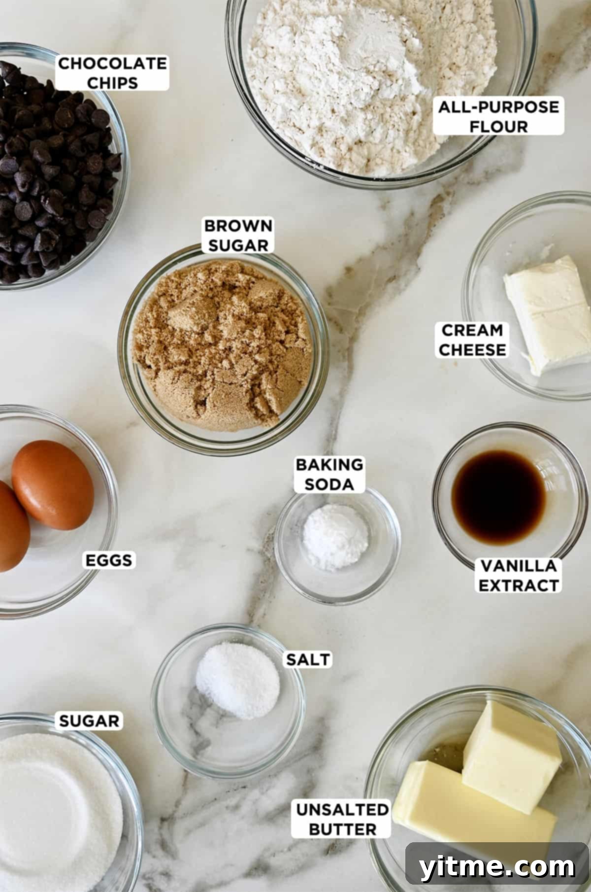 Glass bowls containing ingredients to make soft secret ingredient chocolate chip cookies, including flour, cream cheese, brown sugar, baking soda, vanilla extract, butter, salt, eggs and chocolate chips.