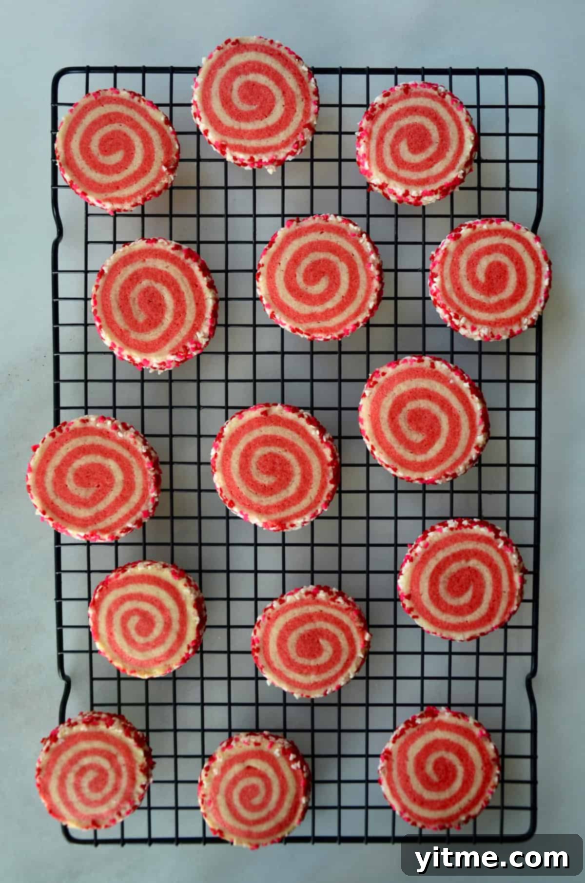 Pink pinwheel sugar cookies, edged in sprinkles, are arranged on a baking rack.