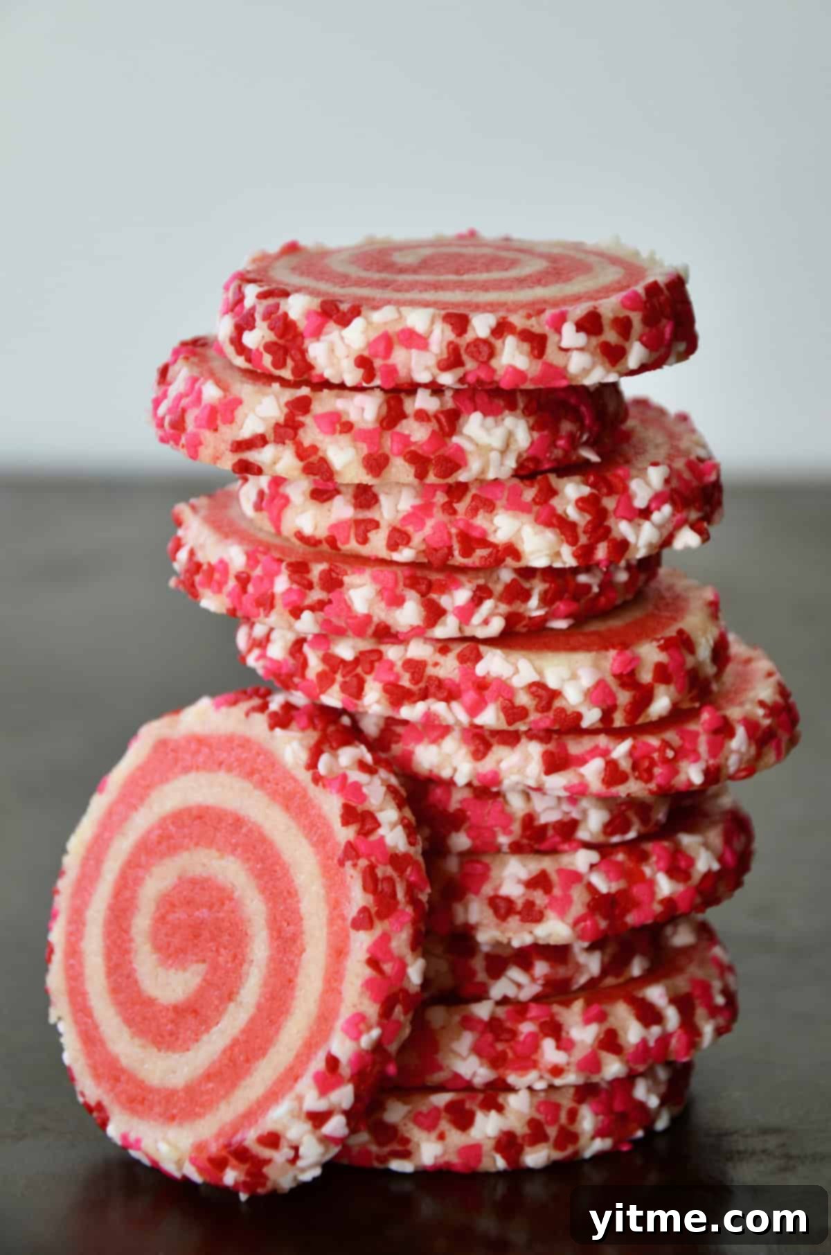 Pink pinwheel sugar cookies, edged in sprinkles, are stacked on a dark surface.