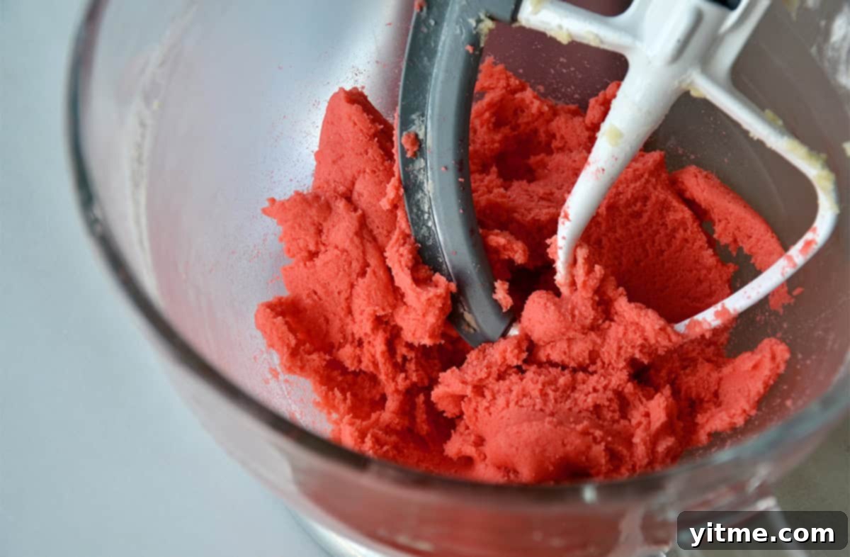 Pink sugar cookie dough is being mixed in a clear glass stand mixer bowl.