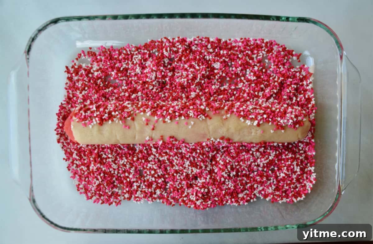 A log of slice and bake sugar cookies is rolled in pink, white and red sprinkles in a clear glass dish.