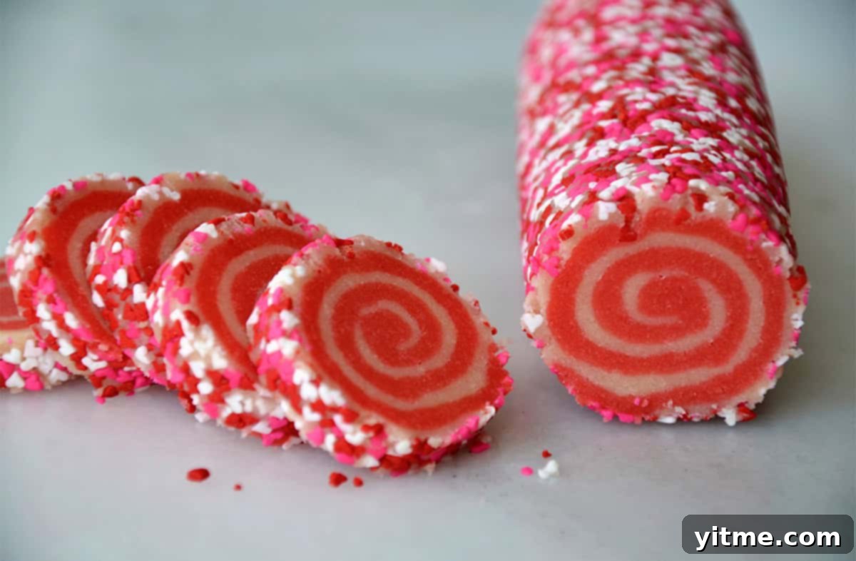 A log of pink and white pinwheel cookie dough, rolled in sprinkles, is cut into rounds.