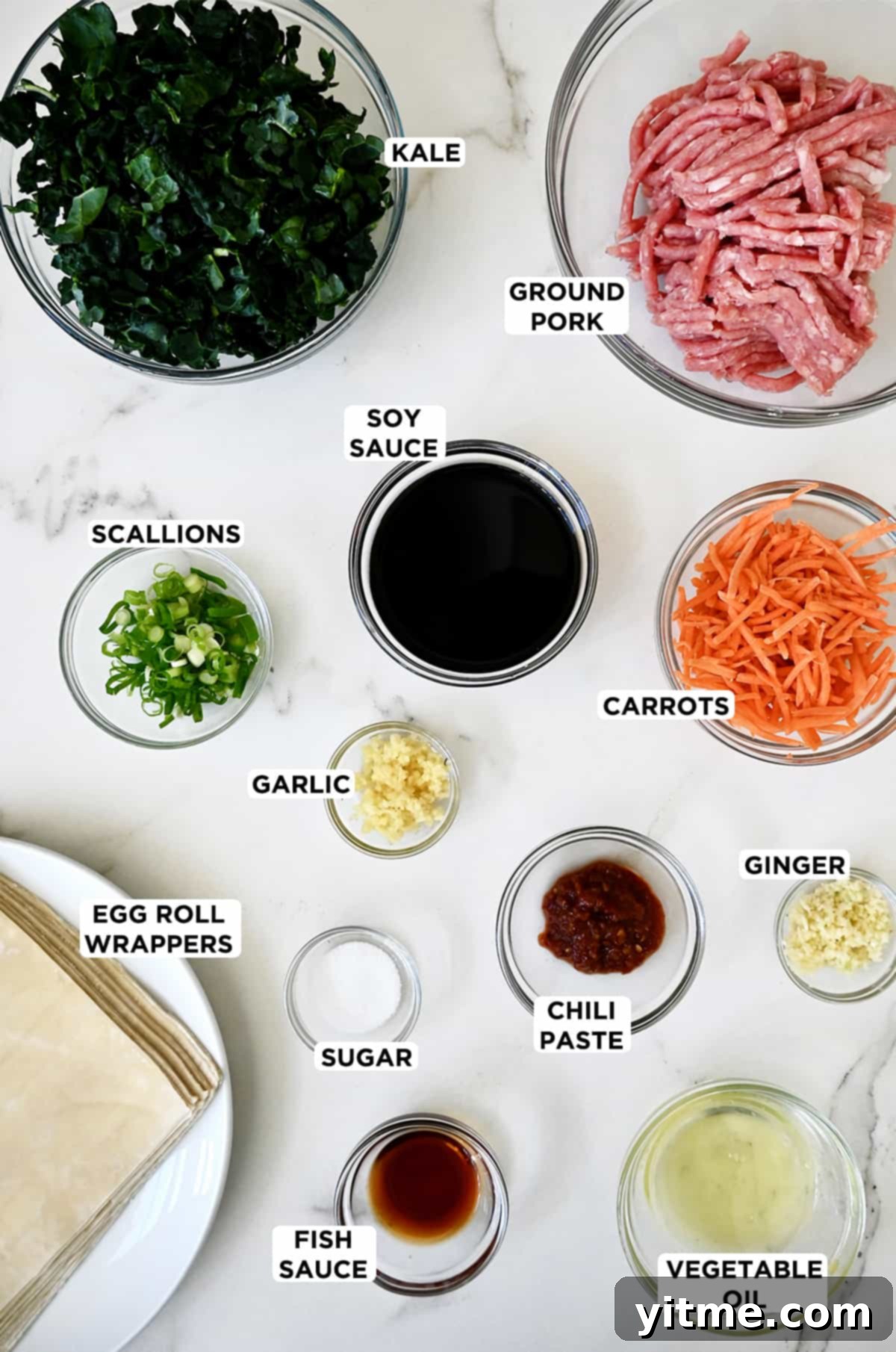 Various sizes of glass bowls containing fresh ingredients for pork egg rolls and ginger soy dipping sauce, including ground pork, vibrant kale, soy sauce, shredded carrots, minced ginger, chili paste, sugar, minced garlic, fresh scallions, fish sauce, vegetable oil, and a stack of egg roll wrappers.