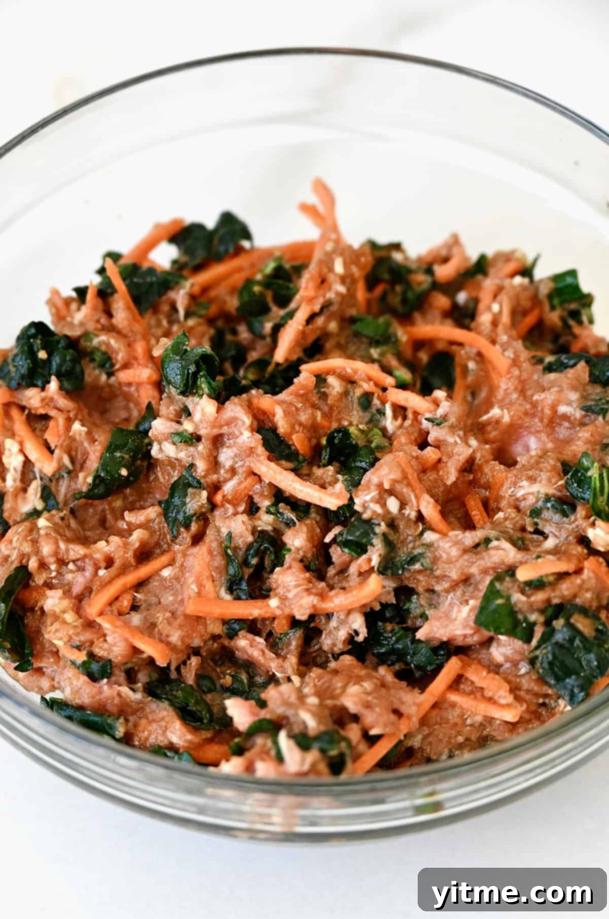 Close-up of a glass bowl containing the vibrant, raw mixture of ground pork, kale, and shredded carrots, ready for filling the egg rolls.