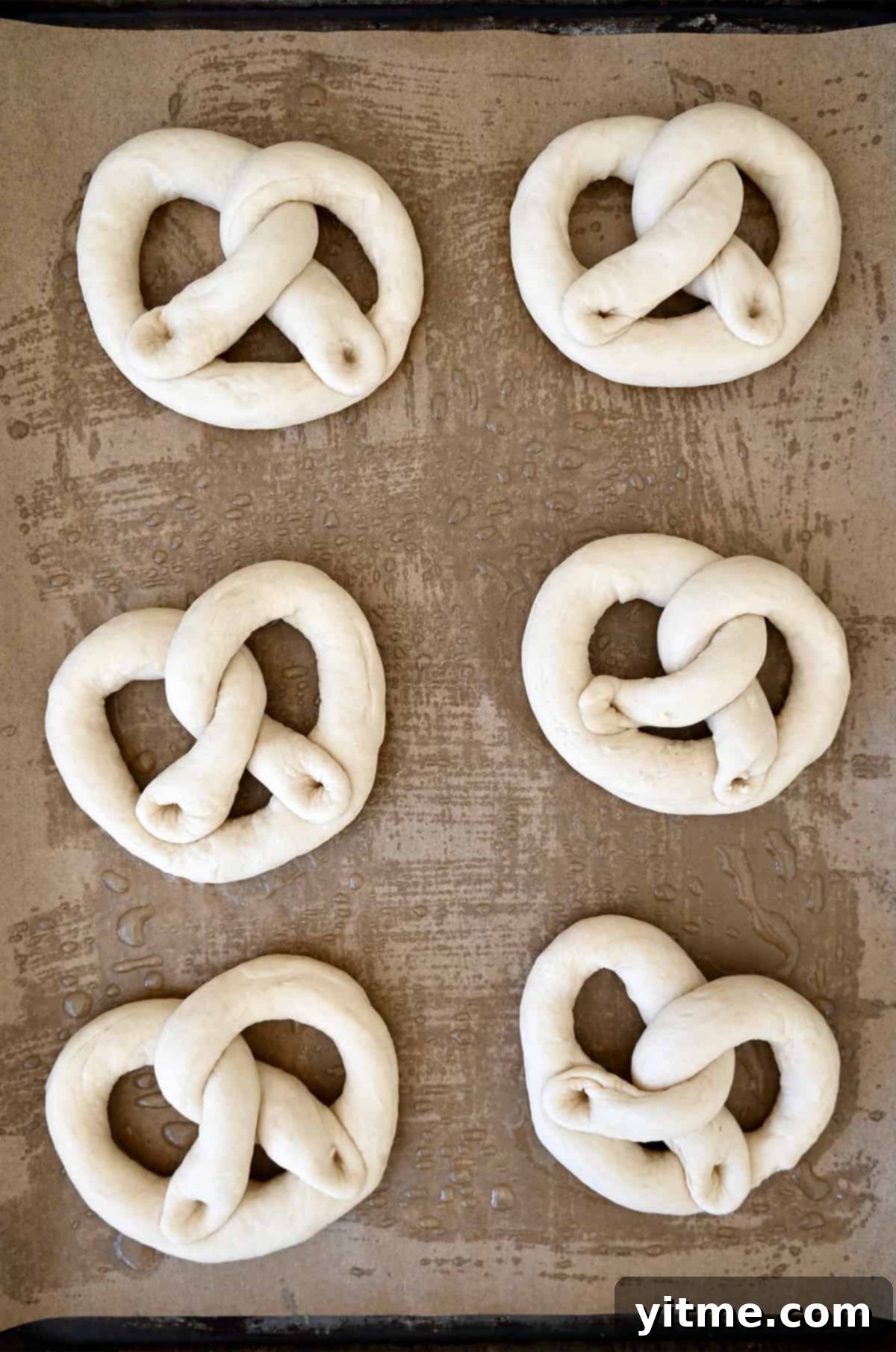 Six iconically-shaped unbaked pretzels on a parchment paper-lined baking sheet.